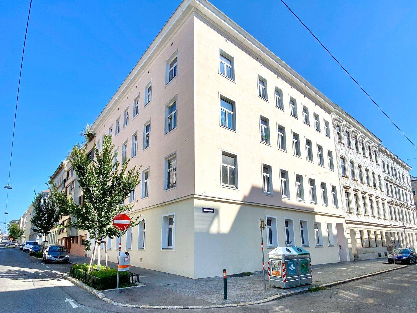 Eckgebäude mit heller Fassade und zahlreichen Fenstern an einer städtischen Straße unter blauem Himmel.