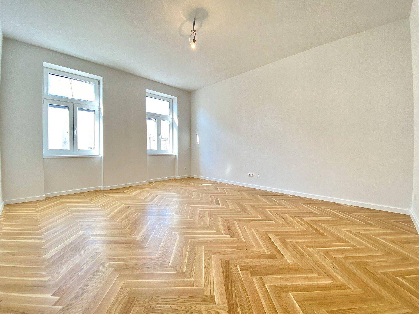 Geräumiges Wohnzimmer mit hellem Fischgrätparkett und zwei großen Fenstern, frisch renoviert.