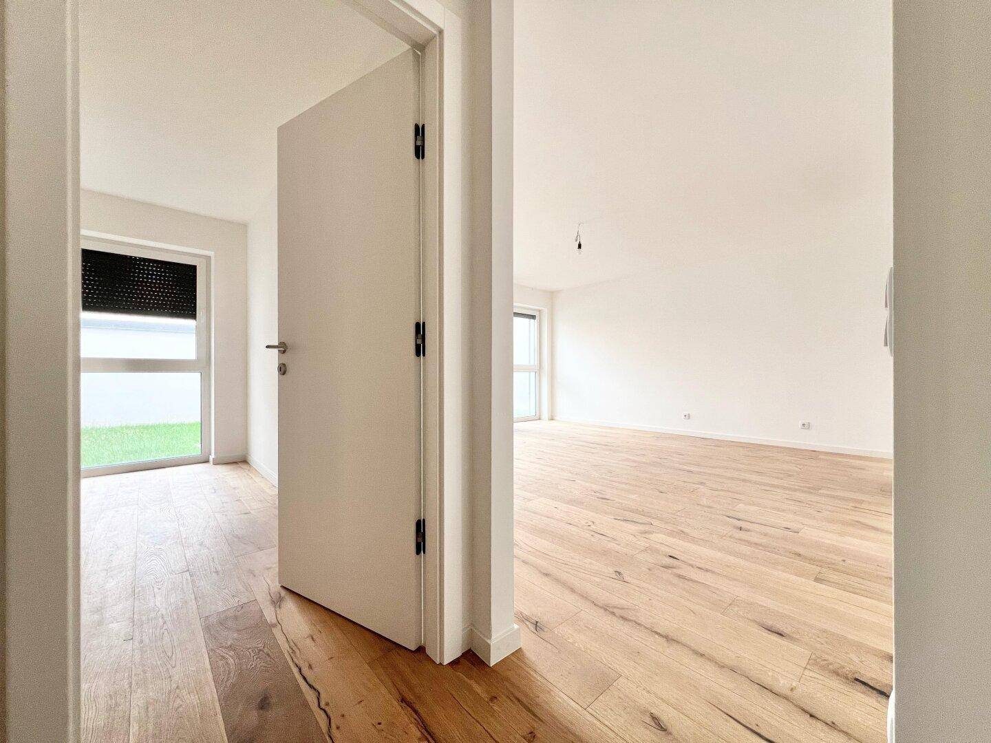 Blick durch eine offene Tür in einen hellen Raum mit Holzfußboden und Fenster mit Rollladen.