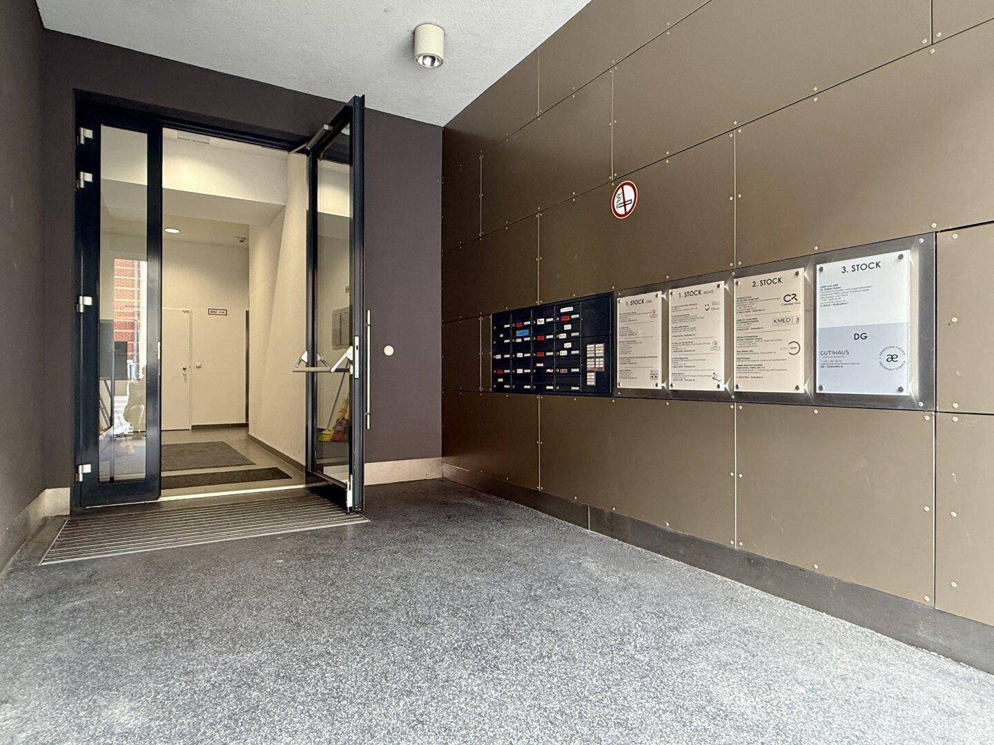 Eingangsbereich eines Gebäudes mit Glastür, Informationsanzeigen und dunklen Wandpaneelen.