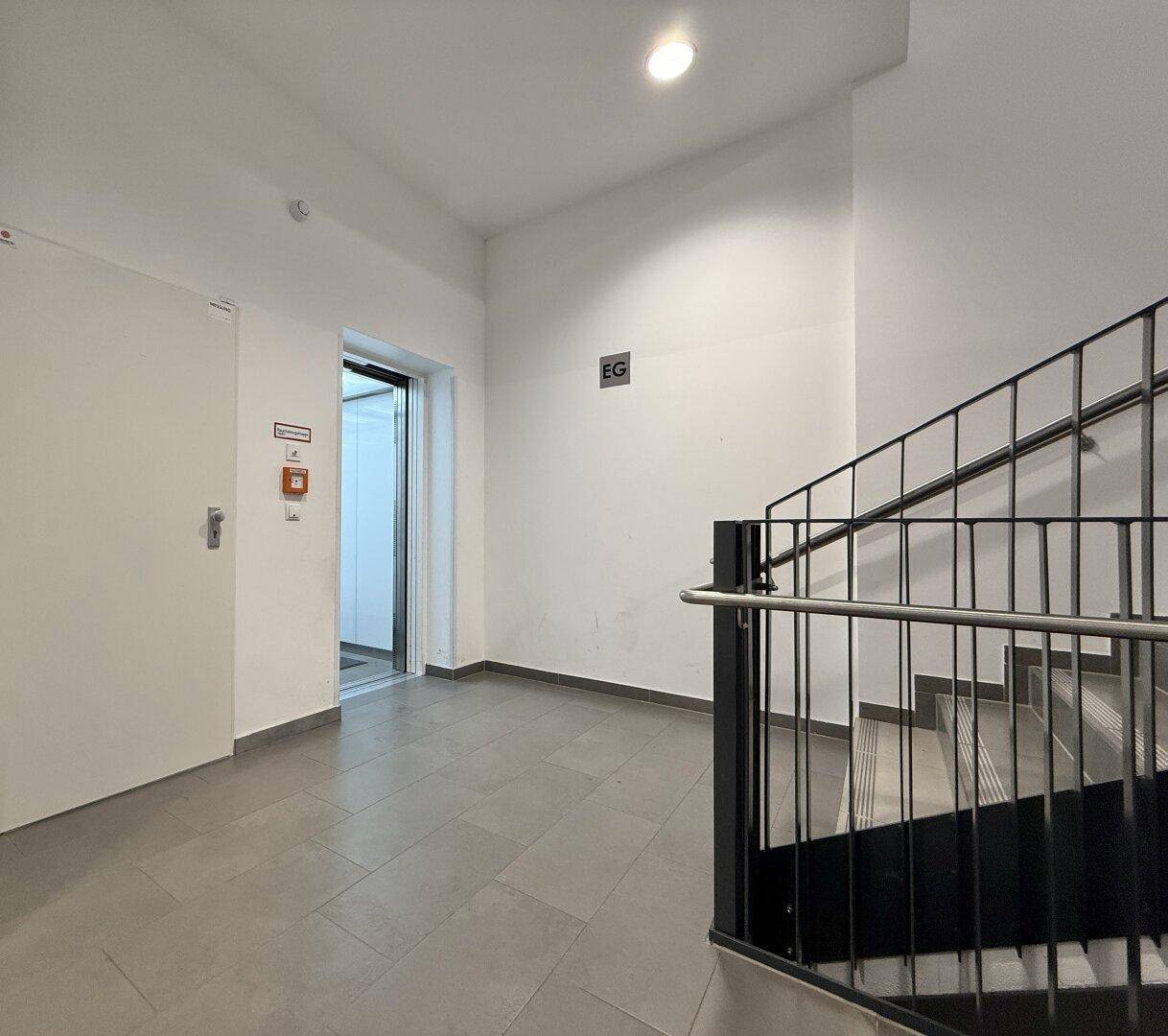 Heller Treppenhausbereich mit Fliesenboden, Metallgeländer und einem sichtbaren Aufzug.