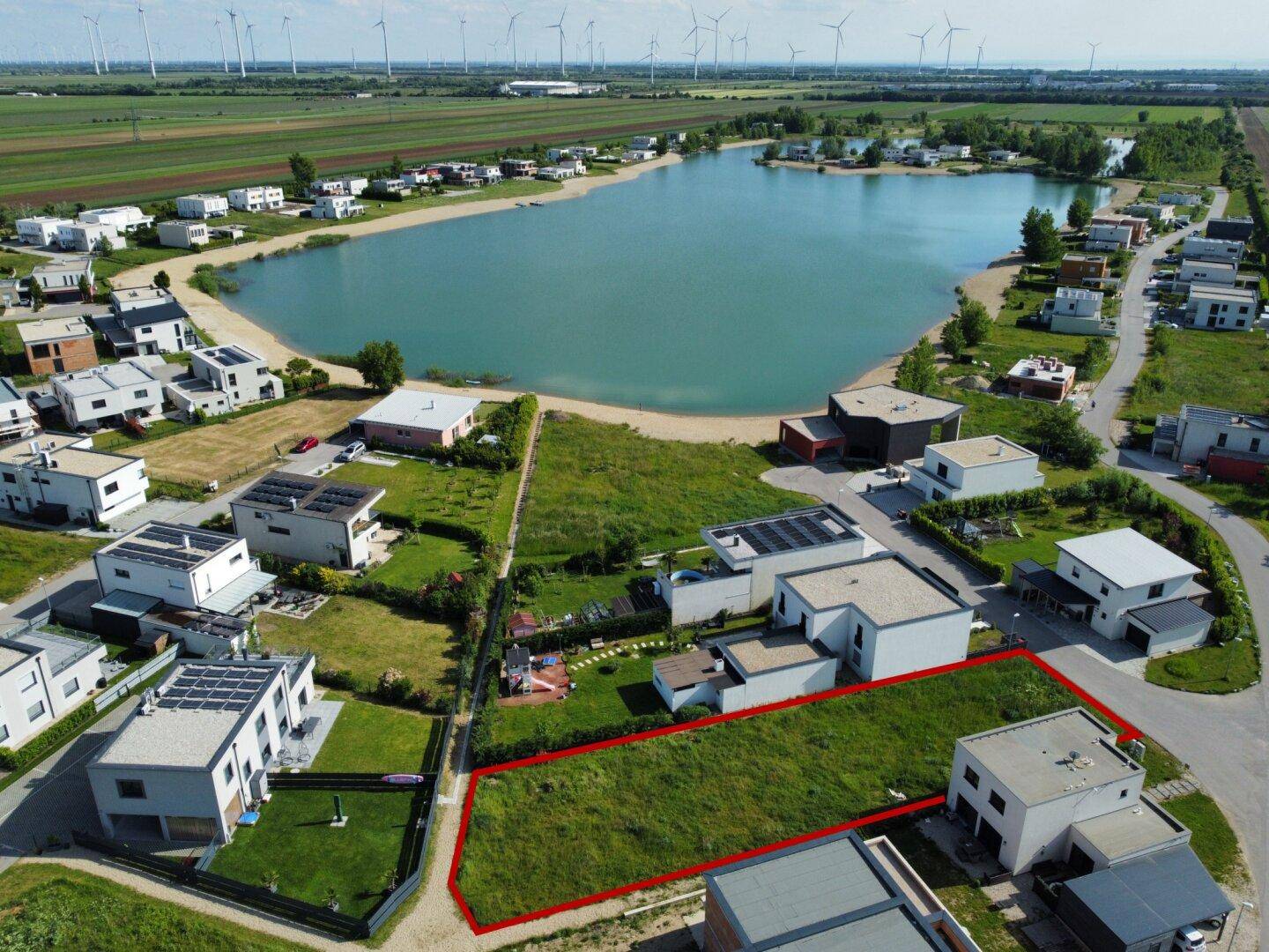 Luftaufnahme eines grünen Baugrundstücks mit modernen Häusern, See und Windkraftanlagen.