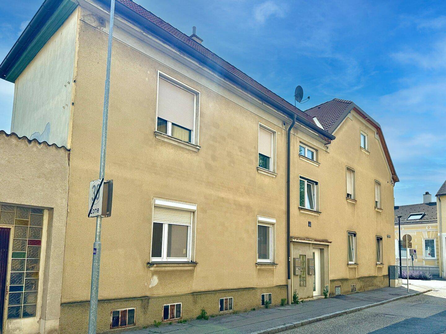 Seitenansicht des Gebäudes mit Eingangsbereich, mehreren Fenstern und Rollläden an der Fassade.