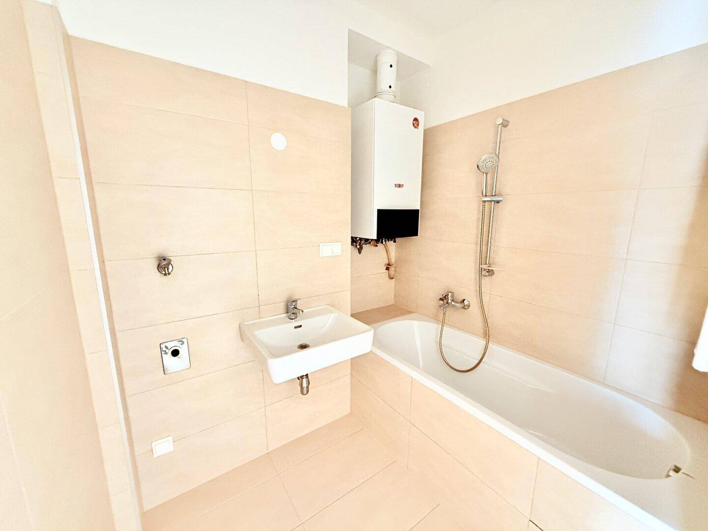 Modern gefliestes Badezimmer mit Badewanne, Waschbecken und Warmwasserbereiter, in neutralen Farbtönen.
