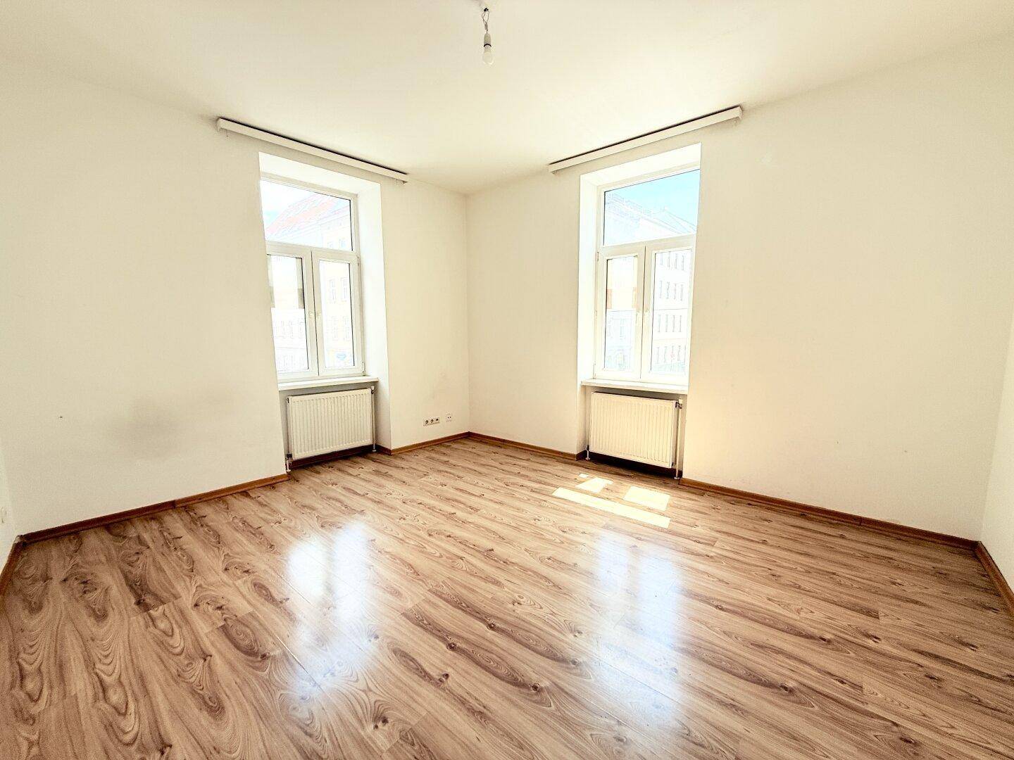 Lichtdurchfluteter Raum mit zwei Fenstern, Parkettboden und Heizkörpern, ideal für Wohn- oder Schlafzimmer.