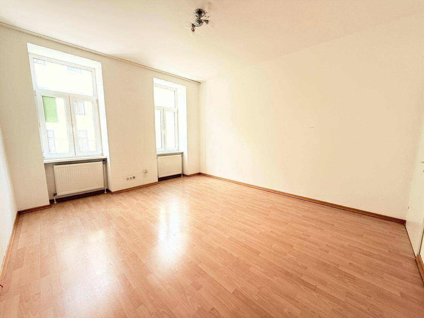 Heller Raum mit zwei Fenstern, Parkettboden und einem Deckenlicht, bietet eine angenehme Atmosphäre.