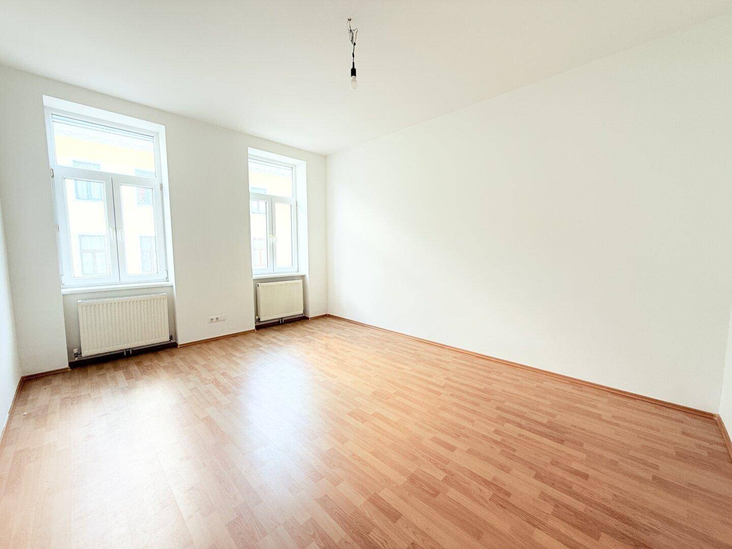 Geräumiger, unmöblierter Raum mit hellem Laminatboden und viel Tageslicht durch zwei Fenster.