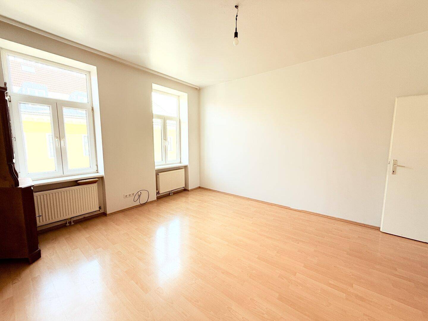 Geräumiger, heller Raum mit Laminatboden, hohen Decken und zwei großen Fenstern.