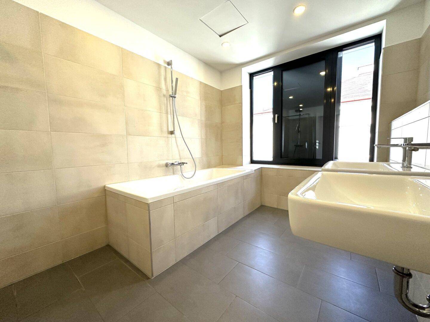 Modernes Badezimmer mit Badewanne, beigefarbenen Fliesen, Doppelwaschbecken und einem Fenster für Tageslicht.