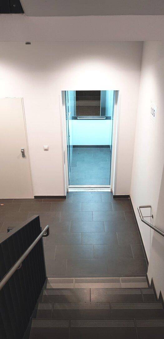 Treppenhaus mit dunklen Fliesen und einem Aufzug im Hintergrund, der durch eine Glastür sichtbar ist.