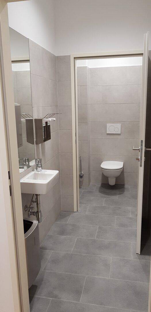 Sauberer Toilettenbereich mit wandhängender Toilette, Waschbecken und großen grauen Wandfliesen.