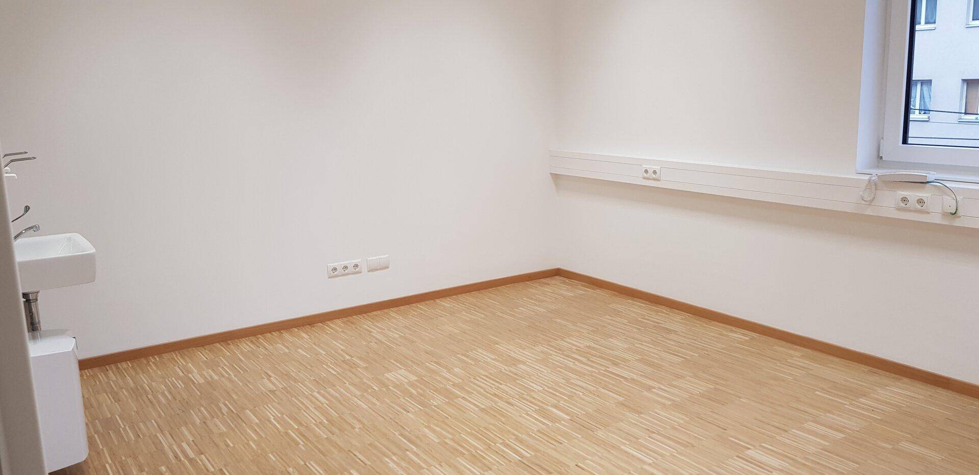 Leerer Raum mit hellem Laminatboden, einem Waschbecken und Steckdosenleisten an der Wand.