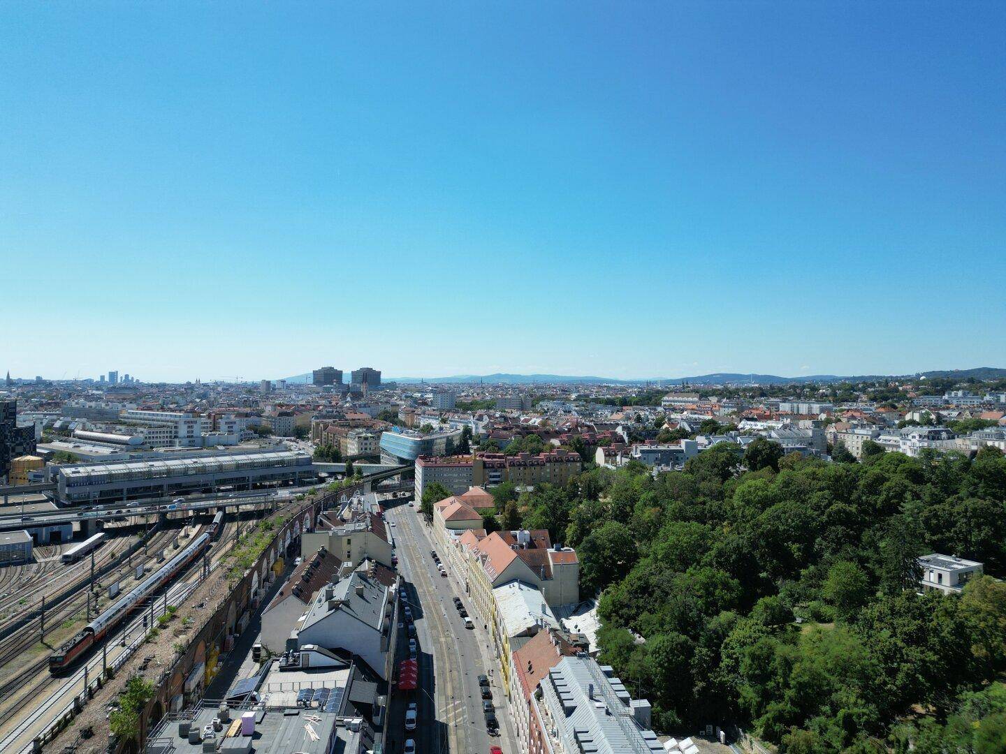 Weitläufige Luftaufnahme einer Stadt mit dichter Bebauung, Grünflächen und einem klaren blauen Himmel.