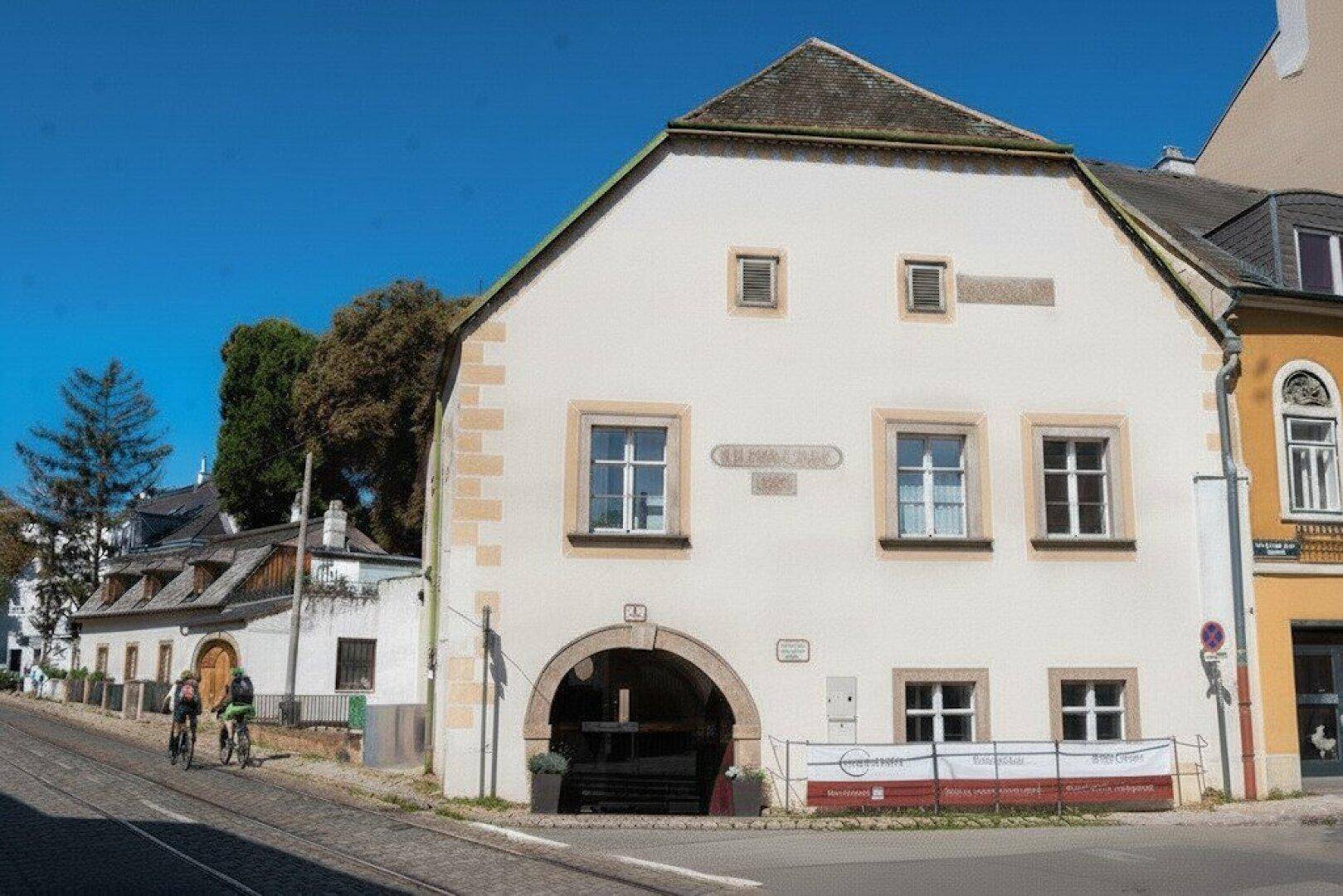 Historisches Gebäude mit weißer Fassade, Bogenportal und traditionellen Fenstern an einer gepflasterten Straße.