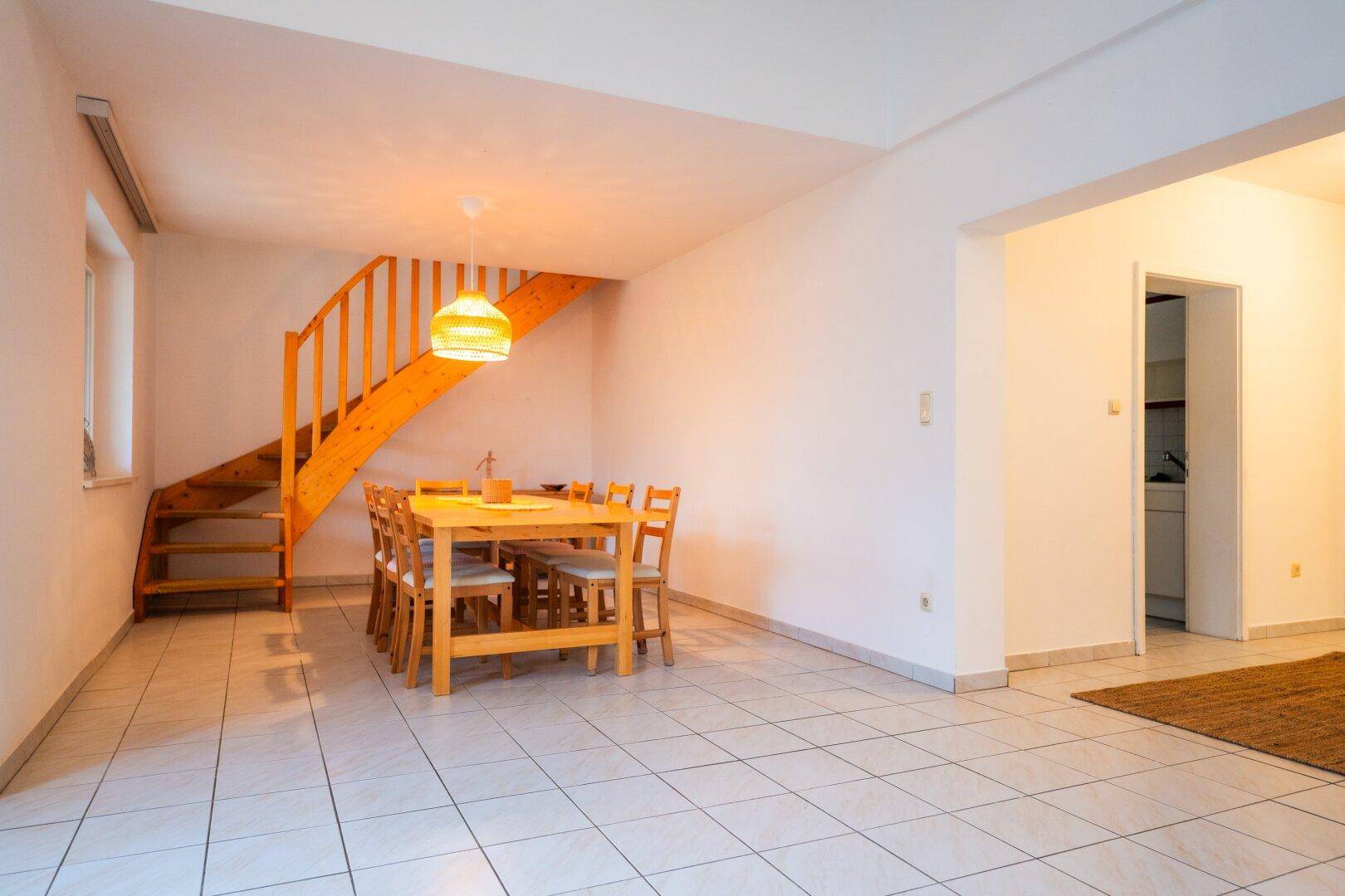 Essbereich mit Holztisch, Stühlen und einer offenen Holztreppe zum oberen Stockwerk.