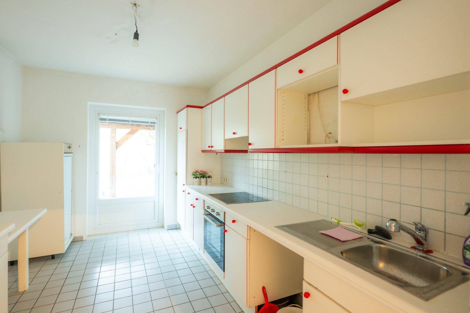 Funktionale Küche mit weißen Schränken, roten Akzenten, Fliesenboden und Fenster.