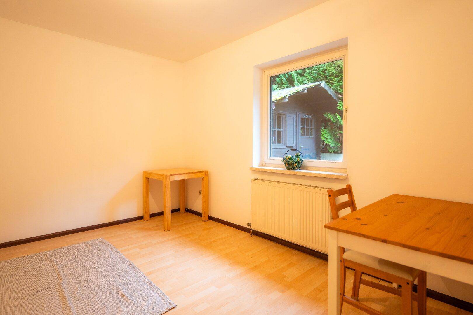 Helles Zimmer mit Holzboden und einem Fenster, das den Blick auf ein kleines Gartenhaus freigibt.