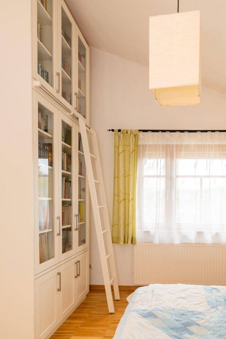 Bücherregal mit Leiter im Schlafzimmer, umgeben von hellen Fenstern und Holzboden.