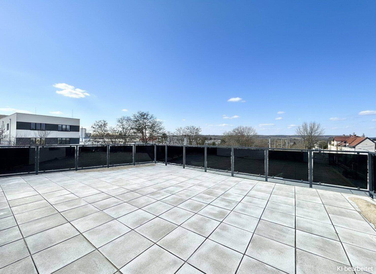 Weitläufige Dachterrasse mit moderner Glasbrüstung und hellem Fliesenboden unter blauem Himmel.