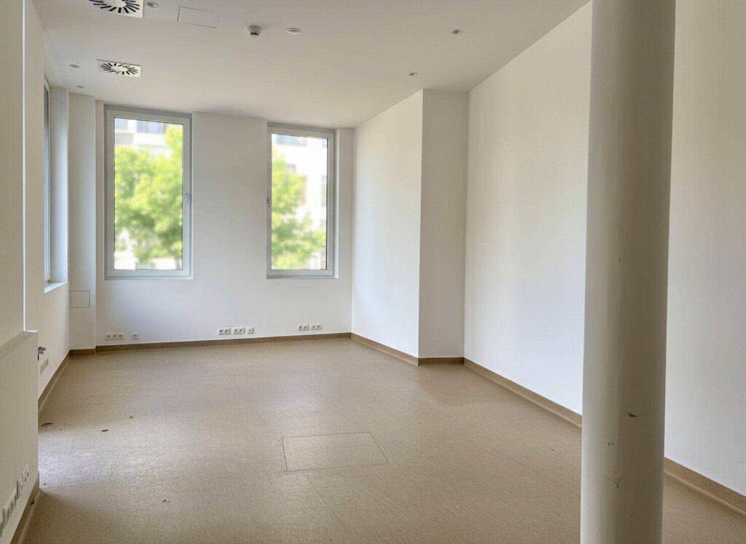 Geräumiger, heller Innenraum mit mehreren Fenstern, einer Säule und Blick ins Grüne.