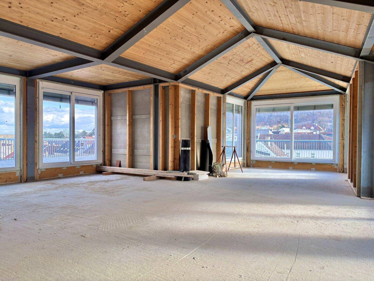 Geräumiger, unfertiger Innenraum mit Holzdachschrägen, großen Fenstern und weitem Blick auf die Umgebung.