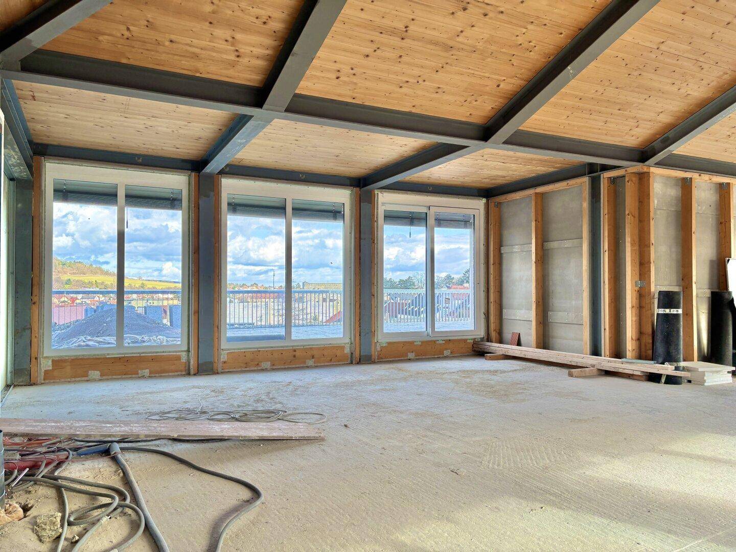 Heller, unfertiger Innenraum mit Holzbalkendecke, großen Fenstern und weitem Ausblick auf die Landschaft.