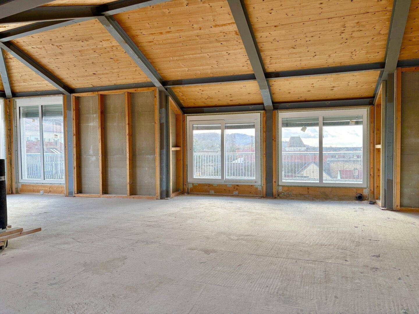 Unfertiger Raum mit Holzdachschrägen, großen Fenstern und Blick auf die Stadt und die umliegenden Hügel.