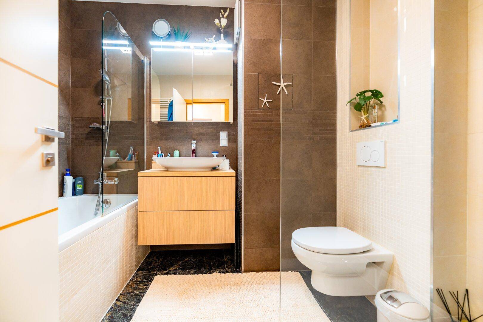 Badezimmer mit Badewanne, Toilette, modernem Waschtisch und braunen Wandfliesen in warmen Tönen.