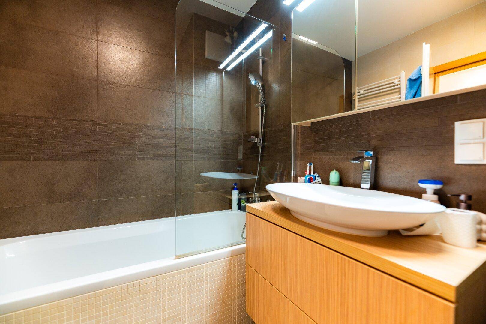 Modernes Badezimmer mit Badewanne, Glasabtrennung, schwebendem Waschtisch und braunen Fliesen.