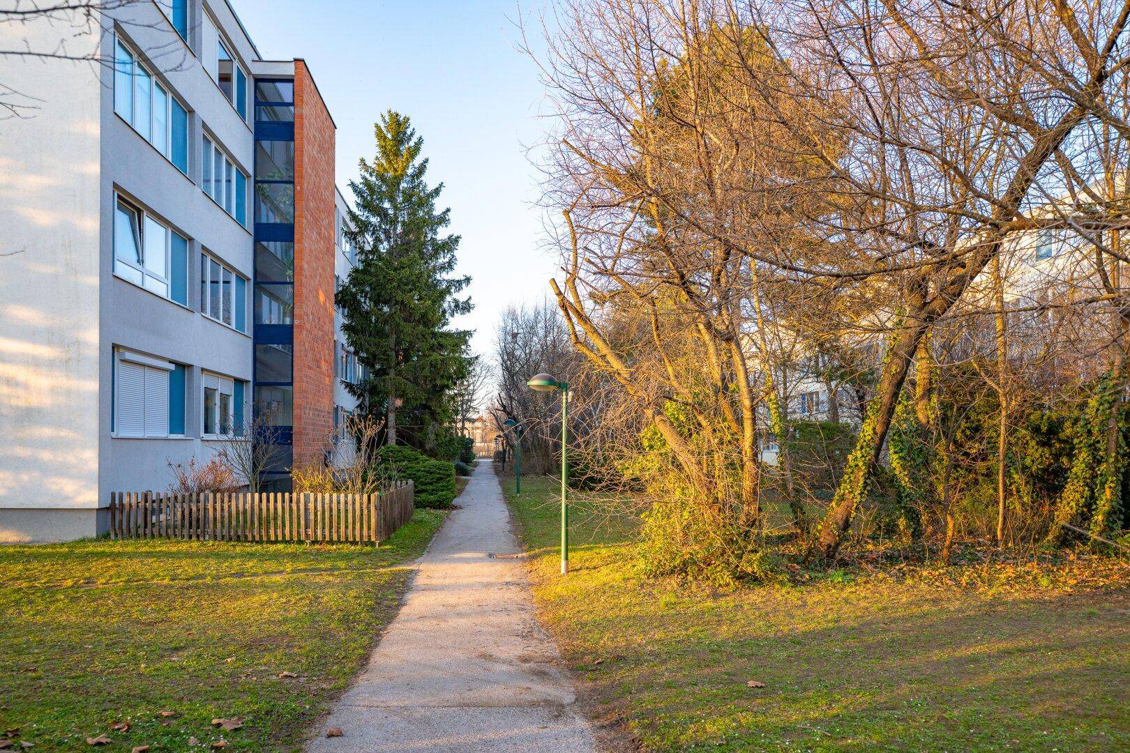Außenansicht eines Wohngebäudes mit Grünflächen und Spazierweg bei Sonnenuntergang.