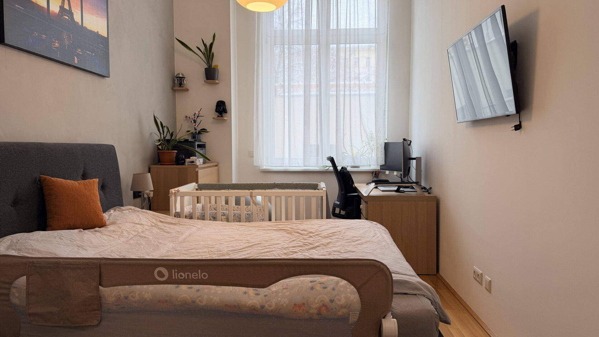 Helles Schlafzimmer mit Doppelbett, Babybett und einem Arbeitsbereich am Fenster.