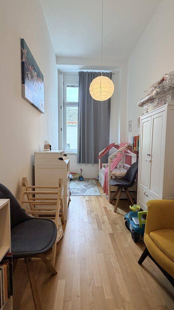 Helles Kinderzimmer mit Holzboden, Babybett und Spielzeug, gut beleuchtet durch ein großes Fenster.