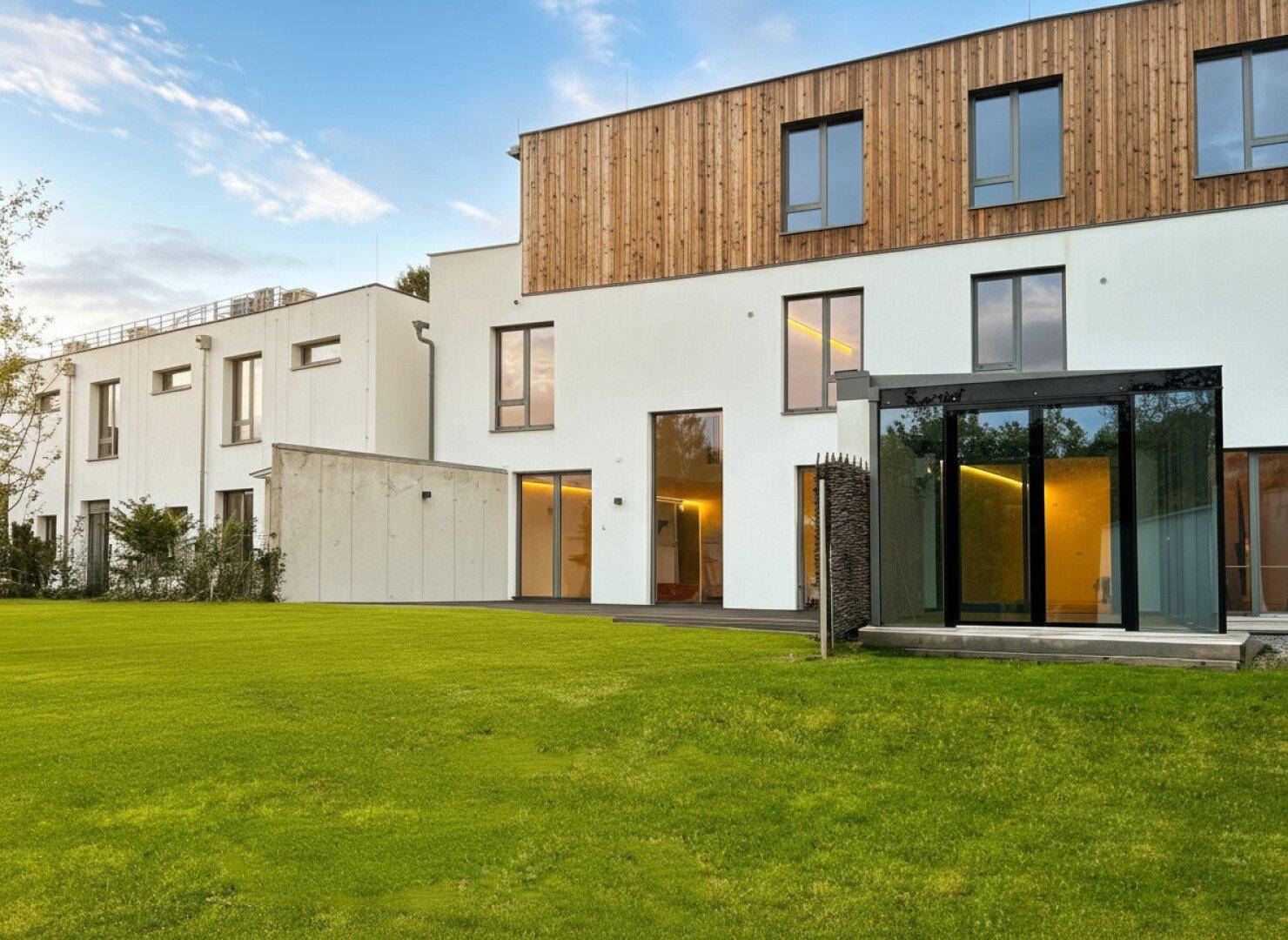 Moderne Architektur mit weißer Fassade, Holzverkleidung und großen Glasfronten, umgeben von einer gepflegten Rasenfläche.