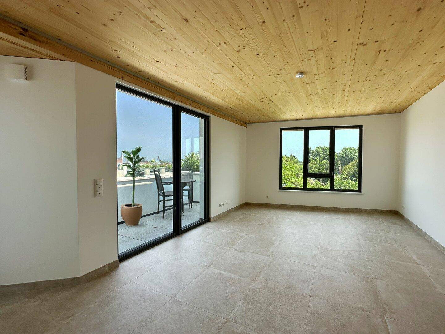 Heller, leerer Raum mit Holzbalkendecke, großen Fenstern und Zugang zu einem Balkon.