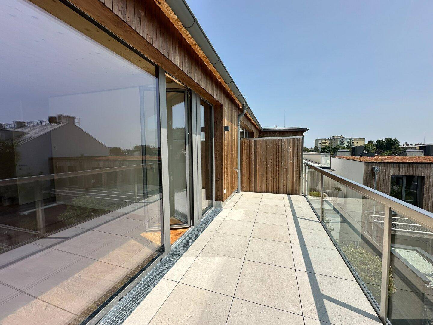 Geräumige Dachterrasse mit Fliesenboden, Glasgeländer und weitem Blick unter blauem Himmel.