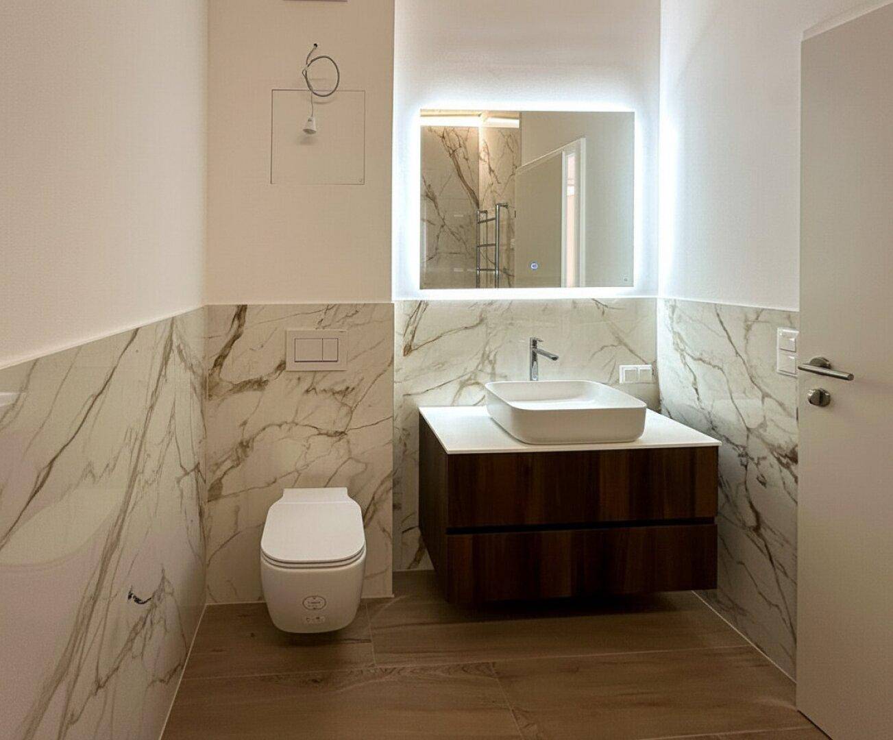 Modernes Badezimmer mit Marmorfliesen, schwebendem Waschtisch und beleuchtetem Spiegel.