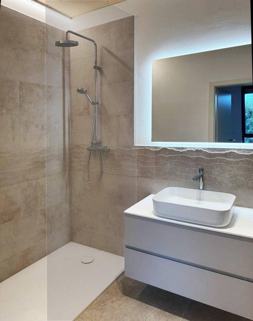 Modernes Badezimmer mit begehbarer Dusche, beigefarbenen Fliesen und beleuchtetem Spiegel.