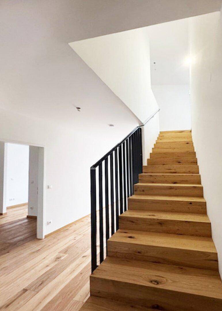 Moderne Holztreppe mit schwarzem Geländer in einem hellen Eingangsbereich.