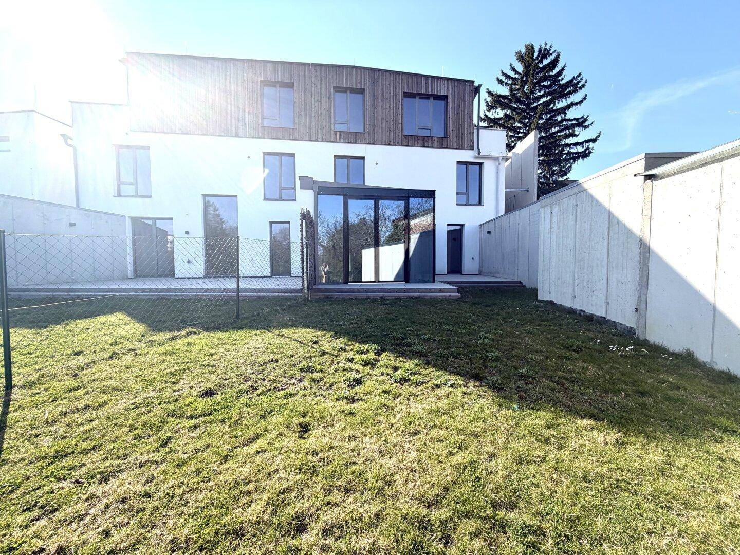 Rückansicht eines modernen Gebäudes mit Wintergartenanbau und sonnigem Gartenbereich.
