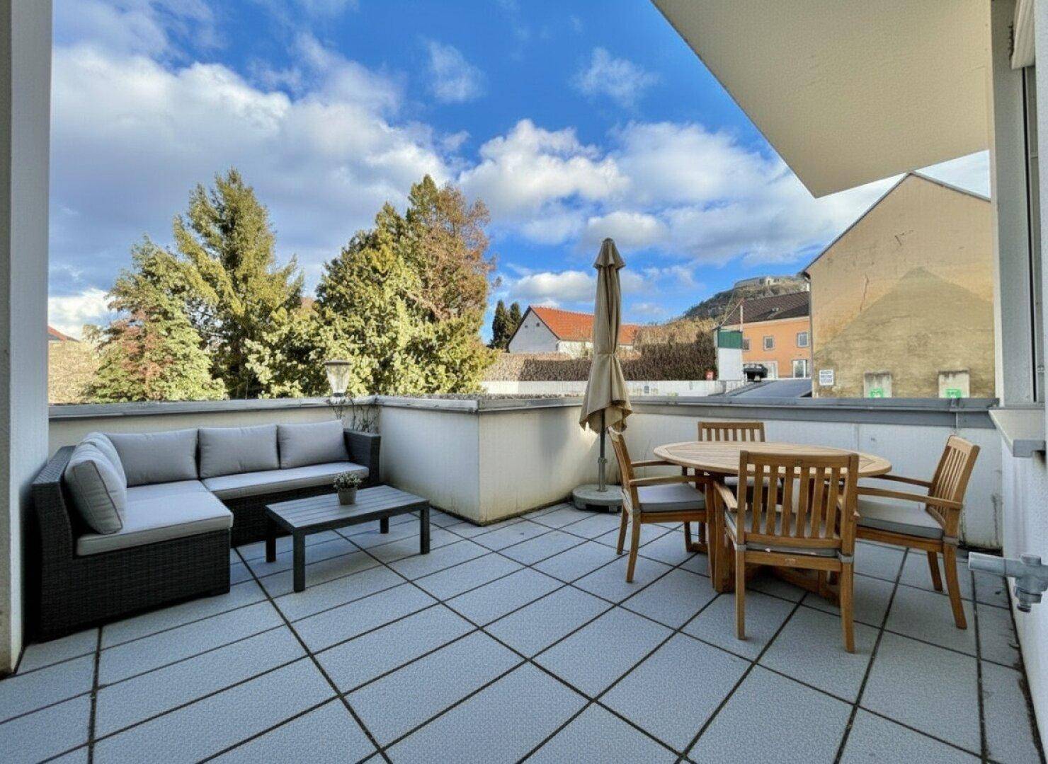 Geräumige Terrasse mit Fliesenboden, bequemen Gartenmöbeln und Blick auf die grüne Umgebung unter blauem Himmel.