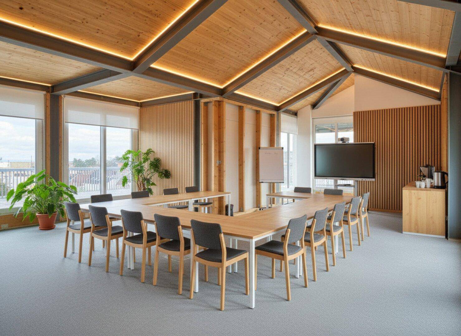 Moderner Konferenzraum mit U-förmigem Tisch, Holzelementen und einem großen Bildschirm für Präsentationen.