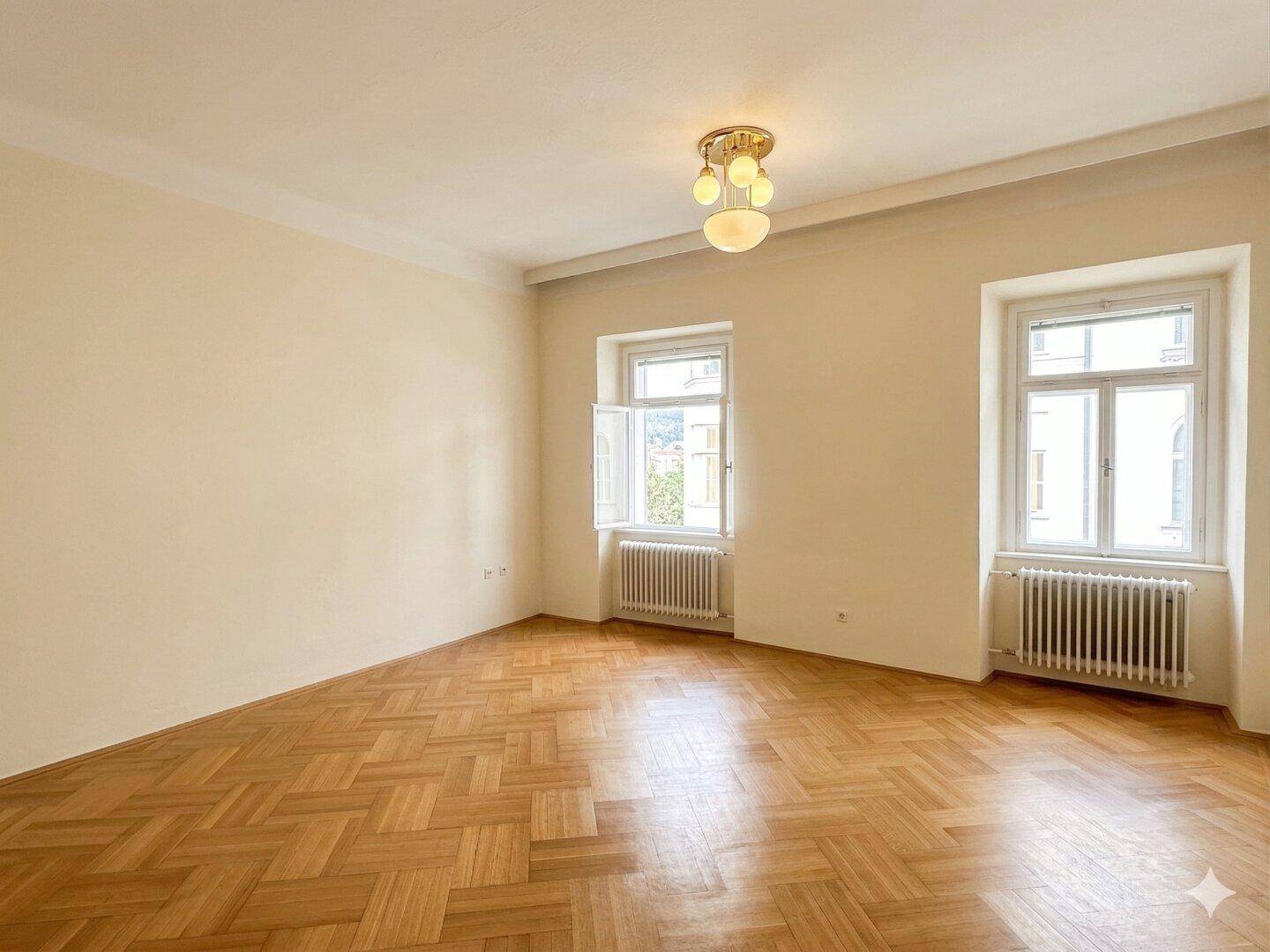 Leerer Raum mit Fischgrätparkett und zwei großen Fenstern, bereit zur individuellen Gestaltung.
