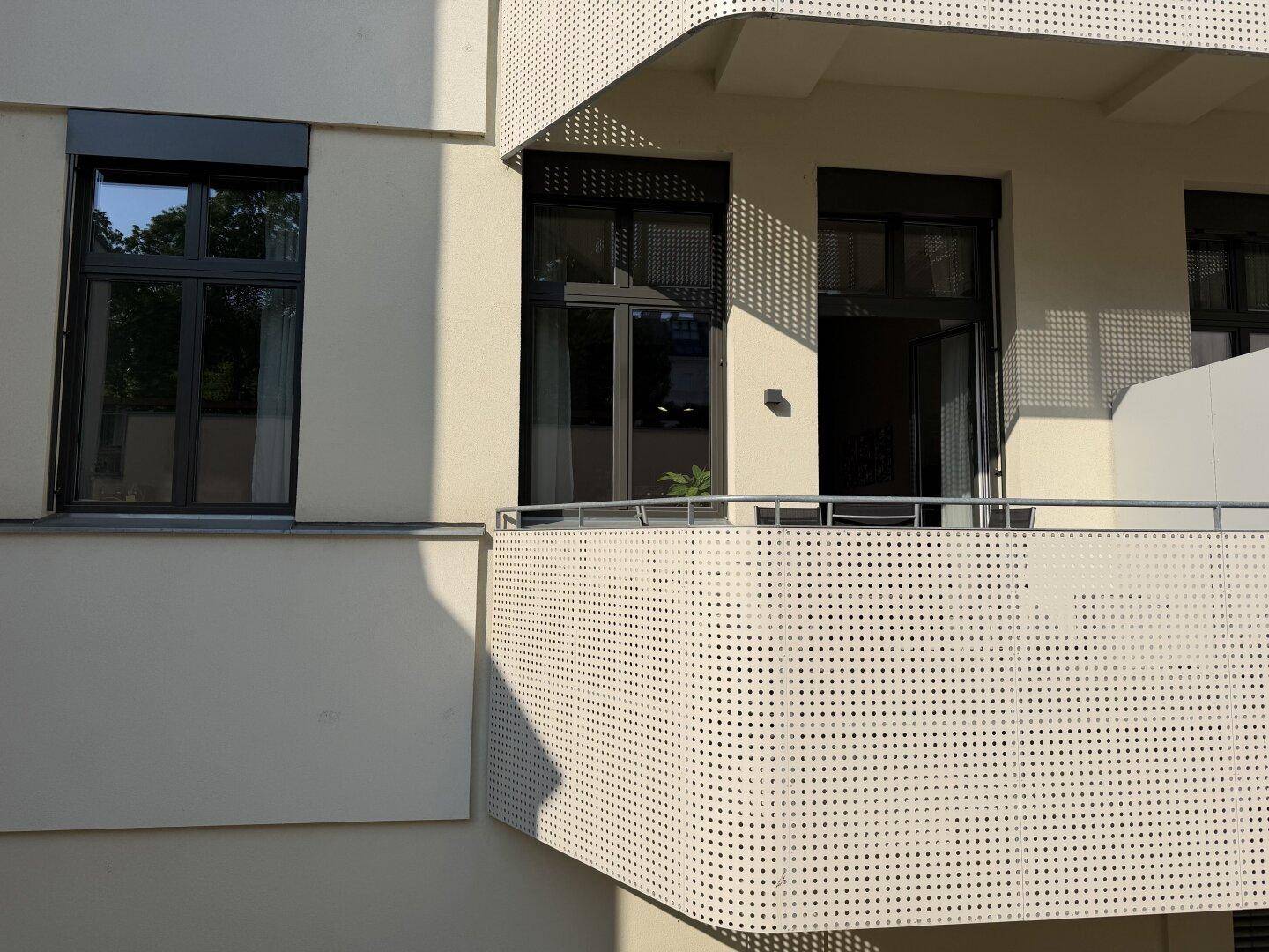 Moderner Balkon mit perforierter Brüstung an einer hellen Gebäudefassade im Sonnenlicht.