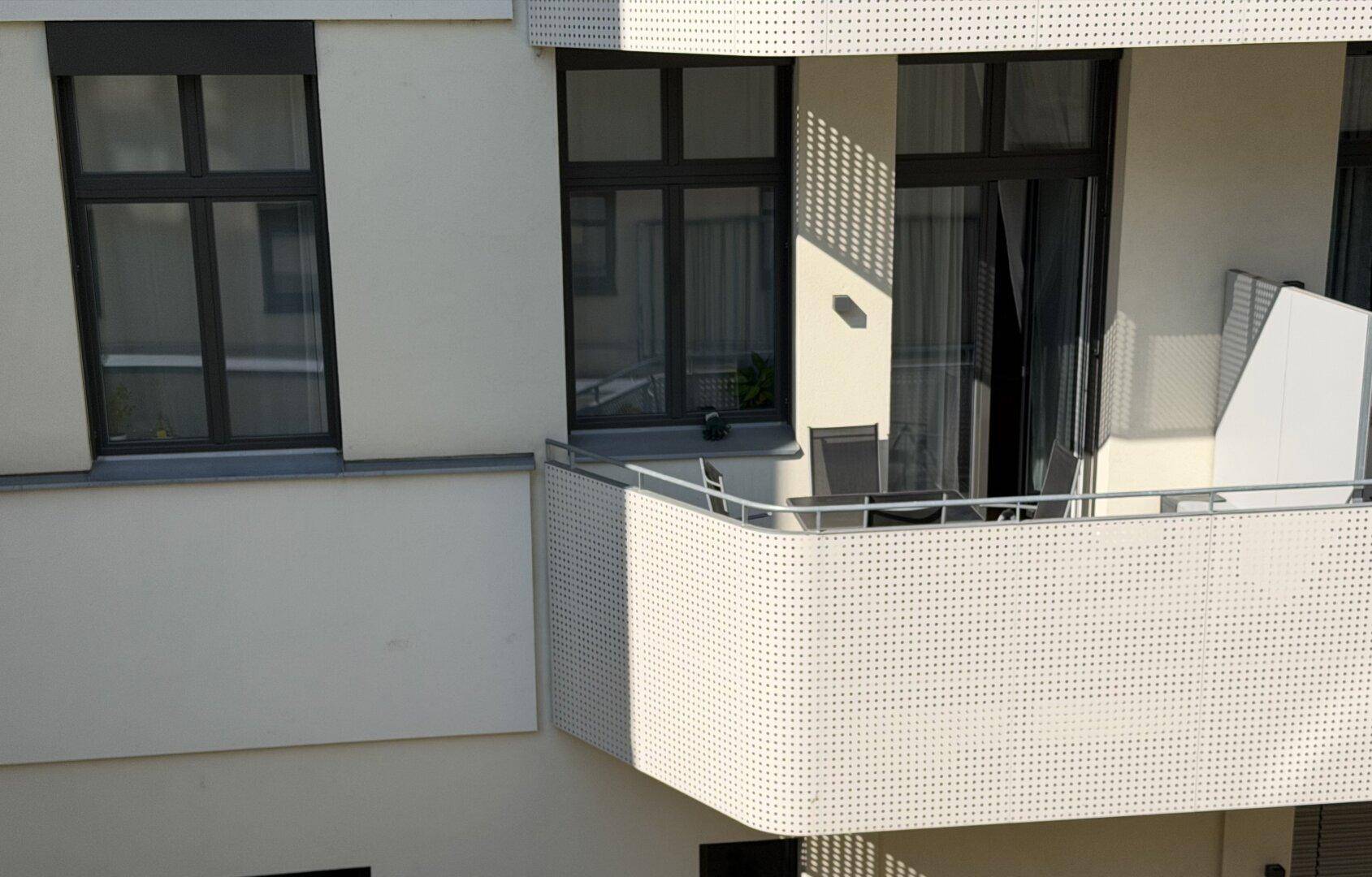 Heller Balkon mit perforierter Brüstung und Sitzgelegenheiten an einer modernen Gebäudefassade.