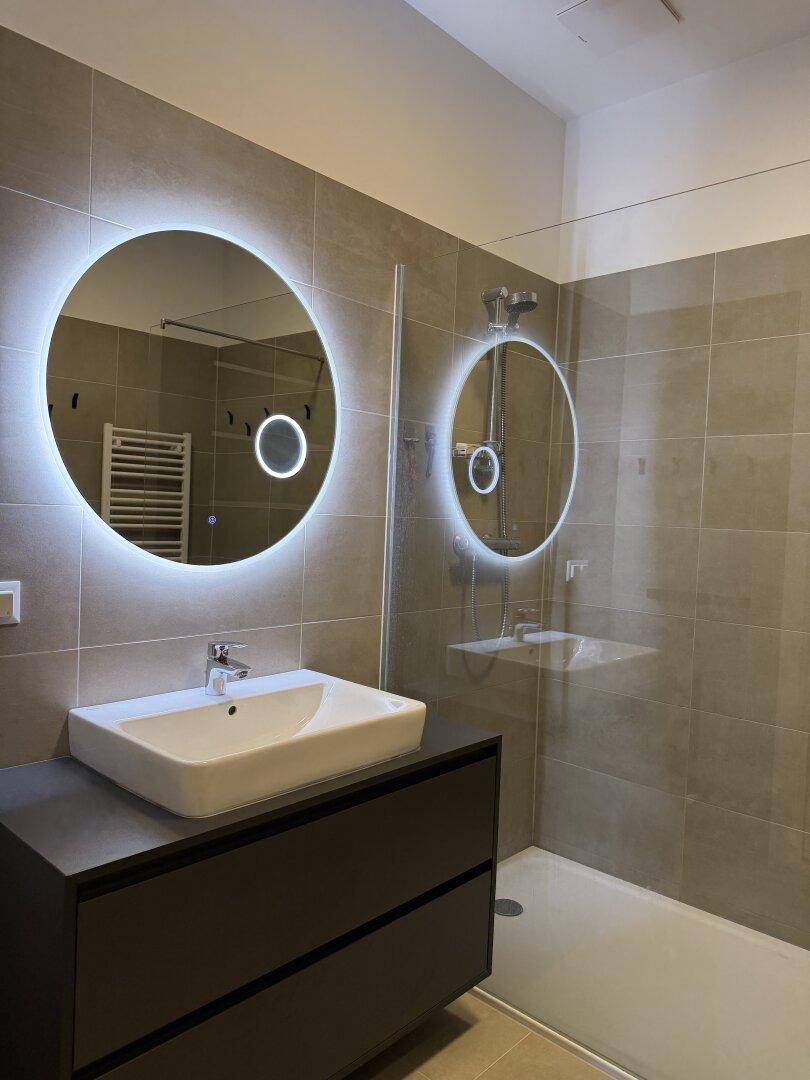 Modernes Badezimmer mit großformatigen Fliesen, beleuchtetem Spiegel und ebenerdiger Dusche.