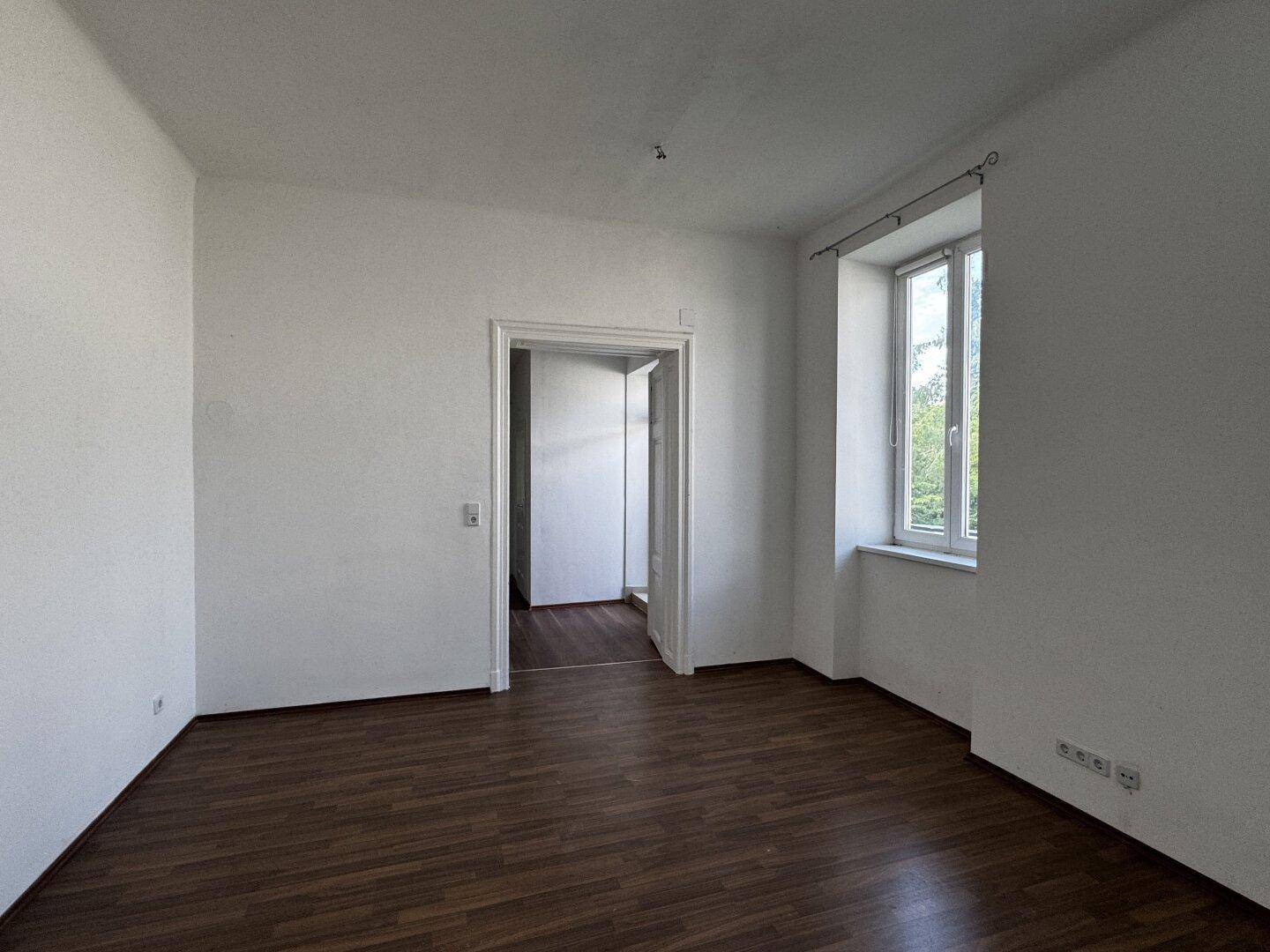 Leerer Raum mit dunklem Holzboden und weißen Wänden, ausgestattet mit einem Fenster und einer Tür.