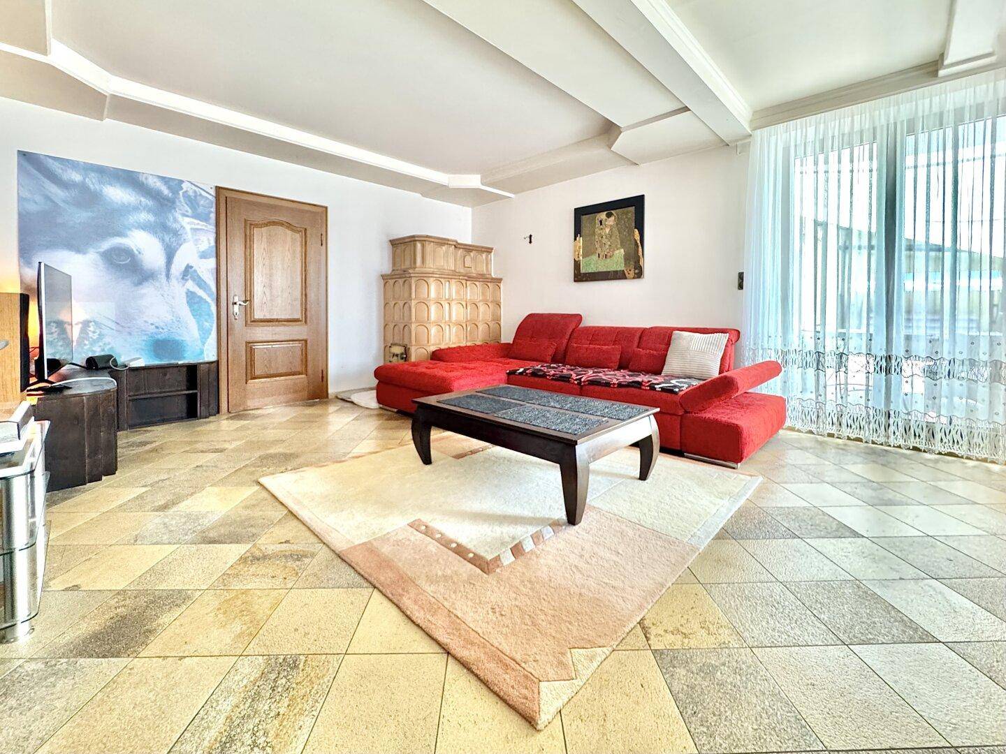 Gemütliches Wohnzimmer mit Kachelofen, rotem Sofa und großen Fenstern.