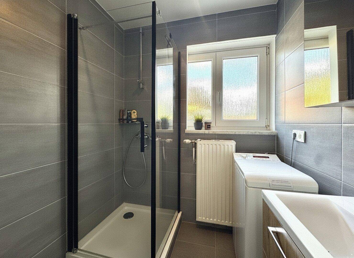 Modernes Badezimmer mit grauen Fliesen, Duschkabine, Waschmaschine und Fenster.