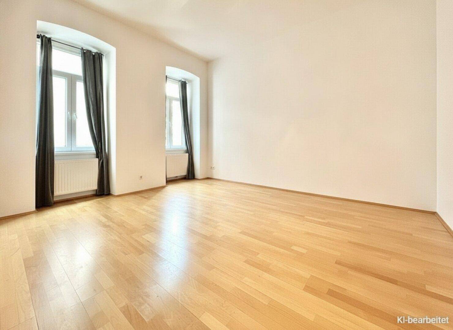 Helles Zimmer mit klassischem Holzparkettboden, weißen Wänden und zwei hohen Fenstern mit dunklen Vorhängen.