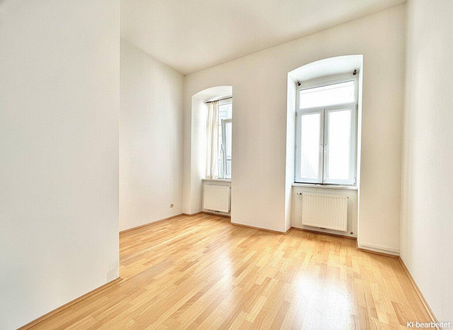 Freundliches, leeres Zimmer mit hellem Holzparkett, weißen Wänden und zwei Fenstern, ideal als Schlaf- oder Arbeitszimmer.